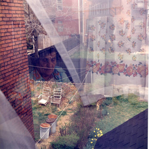 Double image photograph, one layer is of a backyard, the other is an older woman with floral window curtains.